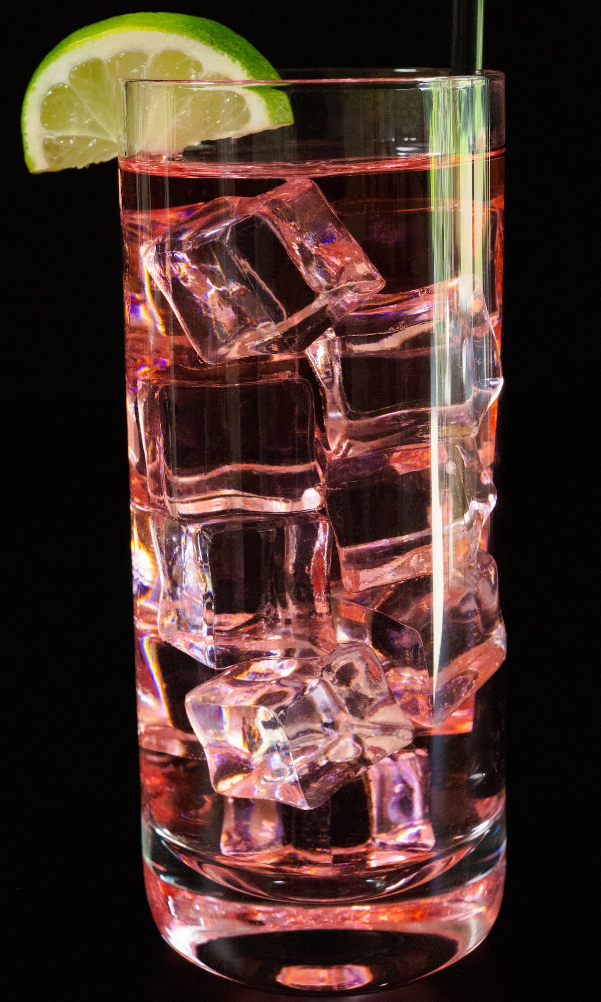 A highball glass is filled with small cubes of ice and a light pink cocktail. It's garnished with a half-moon lime slice and a black straw.
