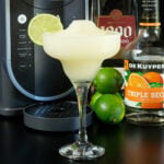 A tall margarita glass filled with a margarita slushie that's garnished with a fresh lime wheel in focus in the foreground, a bottle of tequila, triple sec, a few limes, and a Ninja Slushi machine are out of focus in the background.