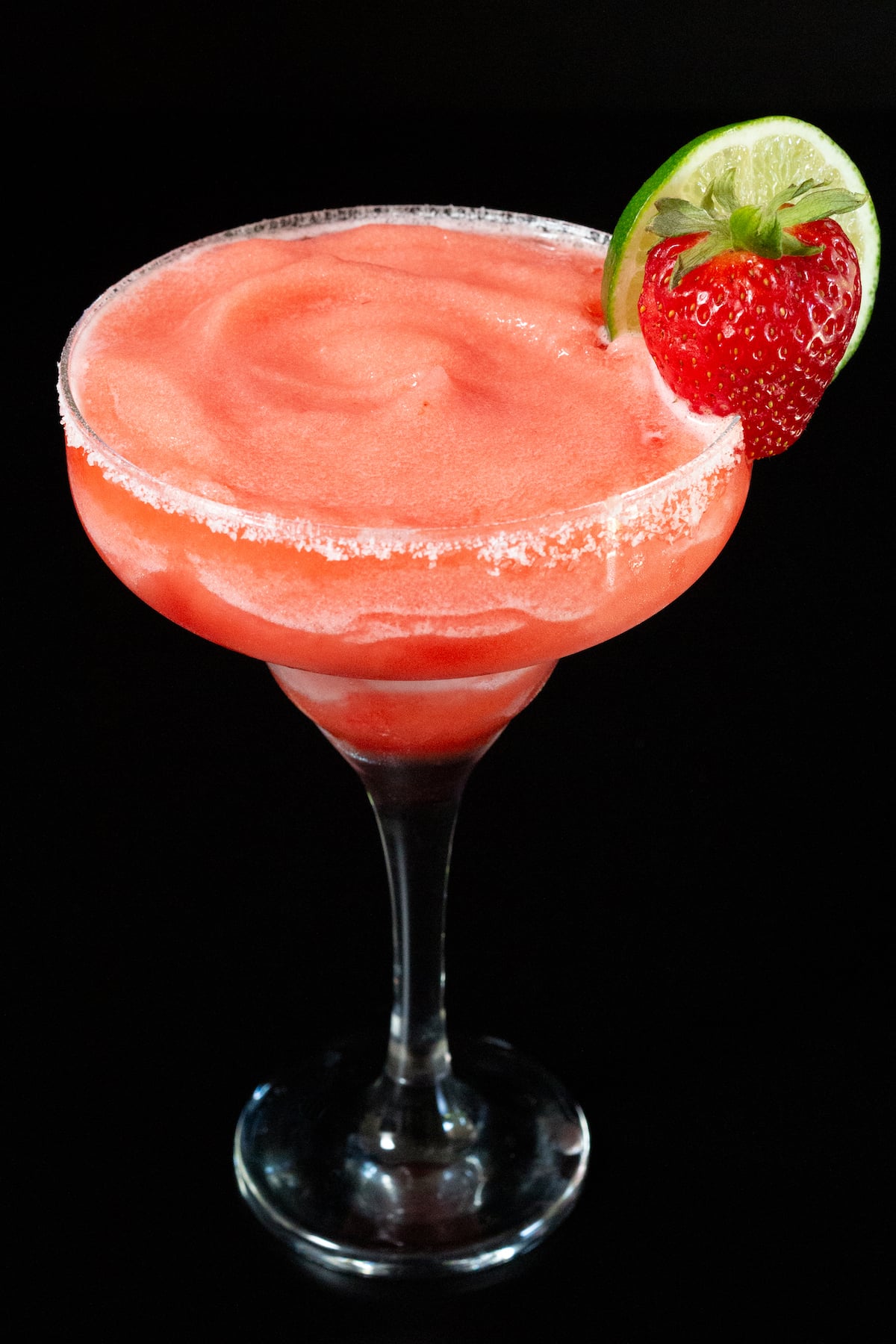 A tall margarita glass rimmed with salt and filled with a frozen strawberry margarita. A fresh lime wheel and whole strawberry are on the right side of the rim as garnish.