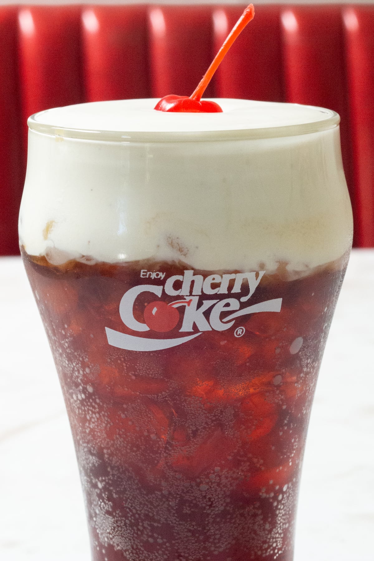 Close up on a pint glass that says "Cherry Coke" and is filled with a reddish brown bubbly soda that's topped with a white vanilla bean cold foam and a maraschino cherry. A red diner booth is out of focus in the background.