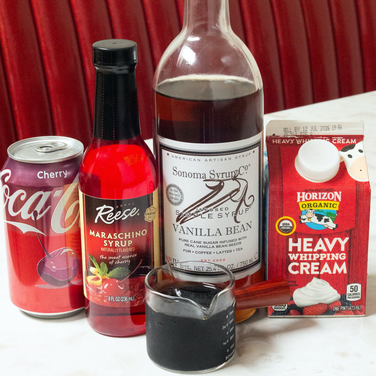 A can of cherry coke, a bottle of maraschino cherry syrup, vanilla bean syrup, a pint of heaving whipping cream, and a double shot of espresso on a diner table.