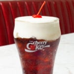 A pint glass with the cherry coke logo is filled with a layered drink that's red at the bottom, reddish-brown in the middle, and topped with a white cold foam and a maraschino cherry. A diner booth is out of focus in the background.