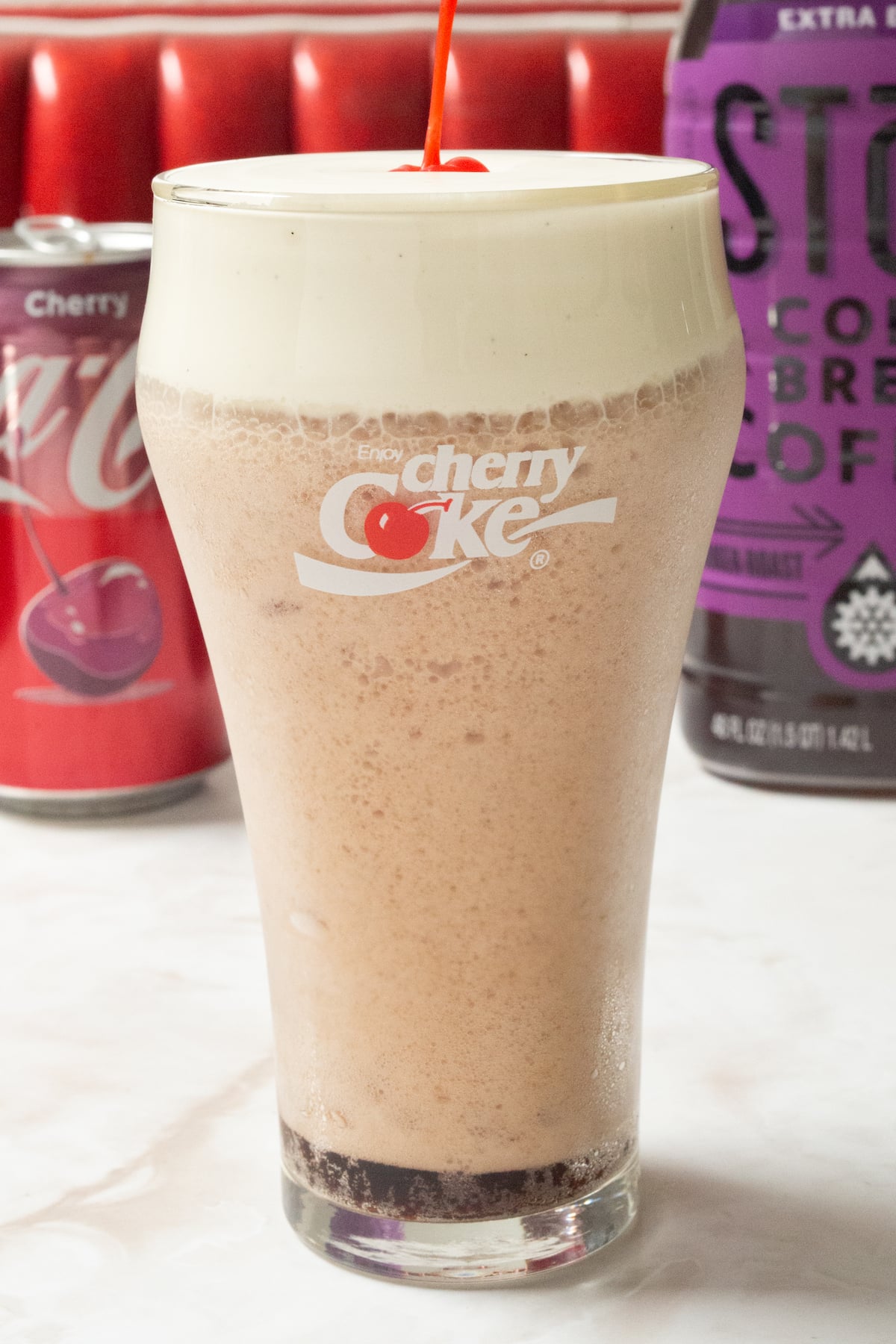 Close up on a pint glass that says "Cherry Coke" and is filled with a reddish brown bubbly soda that's topped with a white vanilla bean cold foam and a maraschino cherry. A red diner booth is out of focus in the background.