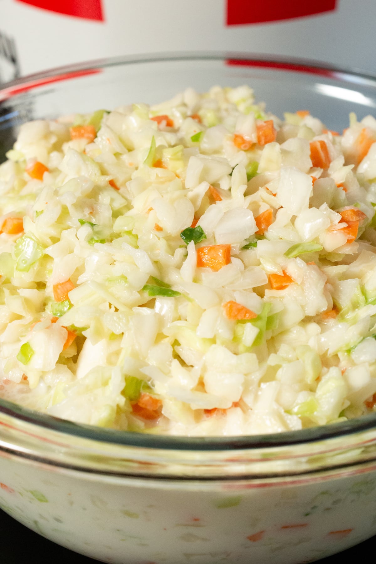A large glass bowl is filled with a homemade version of KFC's coleslaw. It has chopped green cabbage and chopped carrots. A KFC bucket is out of focus in the background.