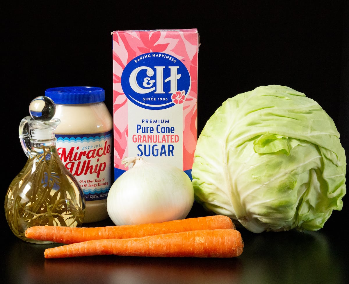 A head of cabbage, two carrots, a white onion, granulated sugar, a jar of miracle whip, and a glass container of tarragon vinegar all on a black background.