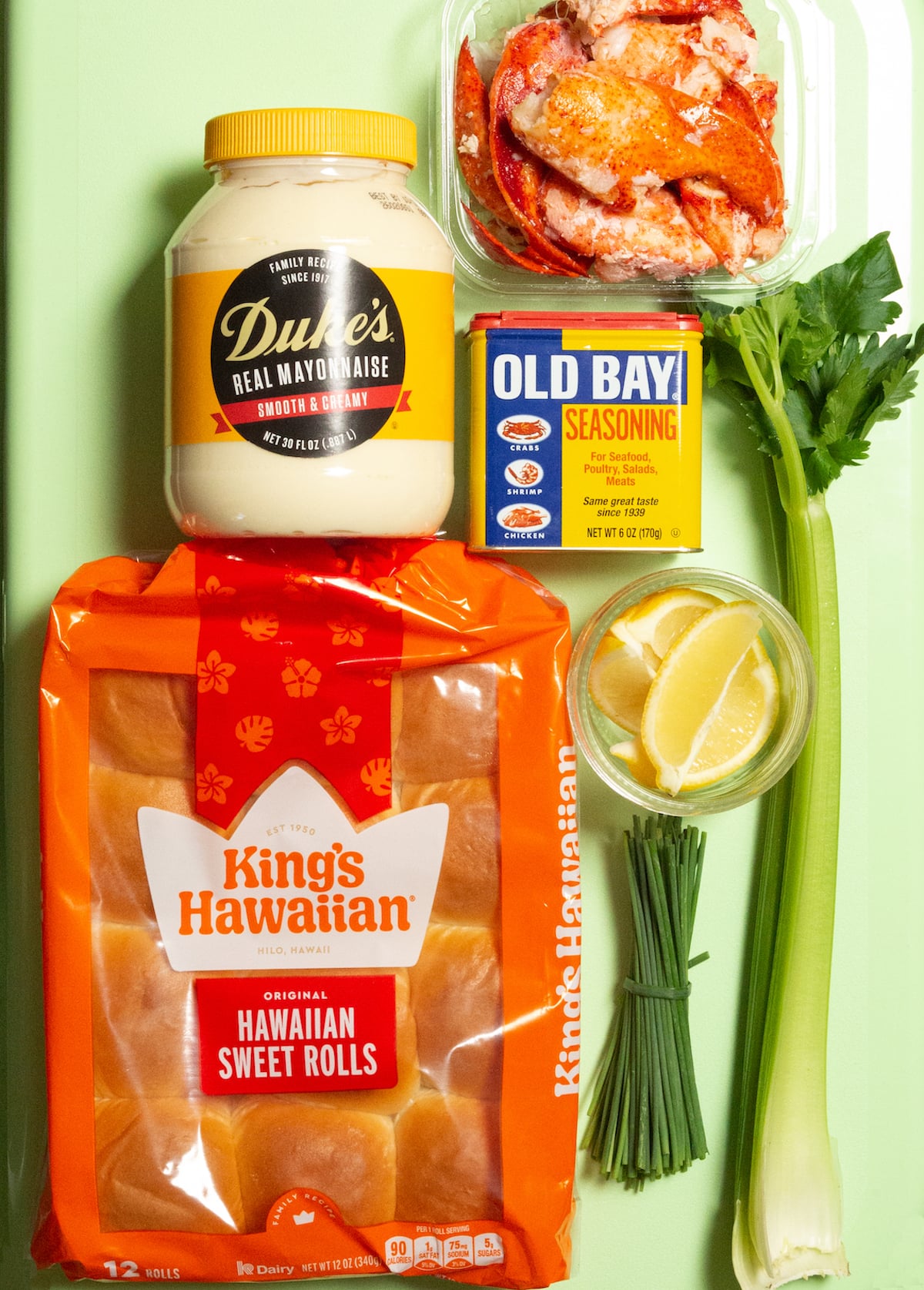 A pack of Hawaiian rolls, a bundle of chives, 2 stalks of celery, lemon wedges, Old bay seasoning, a package of lobster meat, and a container of mayonnaise all on top of a green cooler.