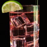 A highball glass is filled with small cubes of ice and a light pink cocktail. It's garnished with a half-moon lime slice and a black straw.