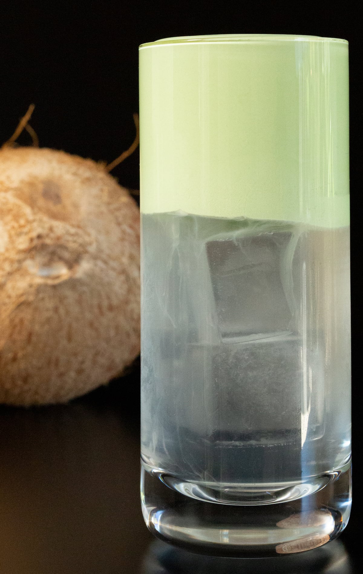 A highball glass half filled with clear block ice cubes and coconut water, and a light green foam at the top. A coconut is out of focus in the background.