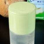 A highball glass is half filled with clear coconut water and the top half is a light green matcha cold foam. A brown coconut and a container of coconut water are out of focus in the background.