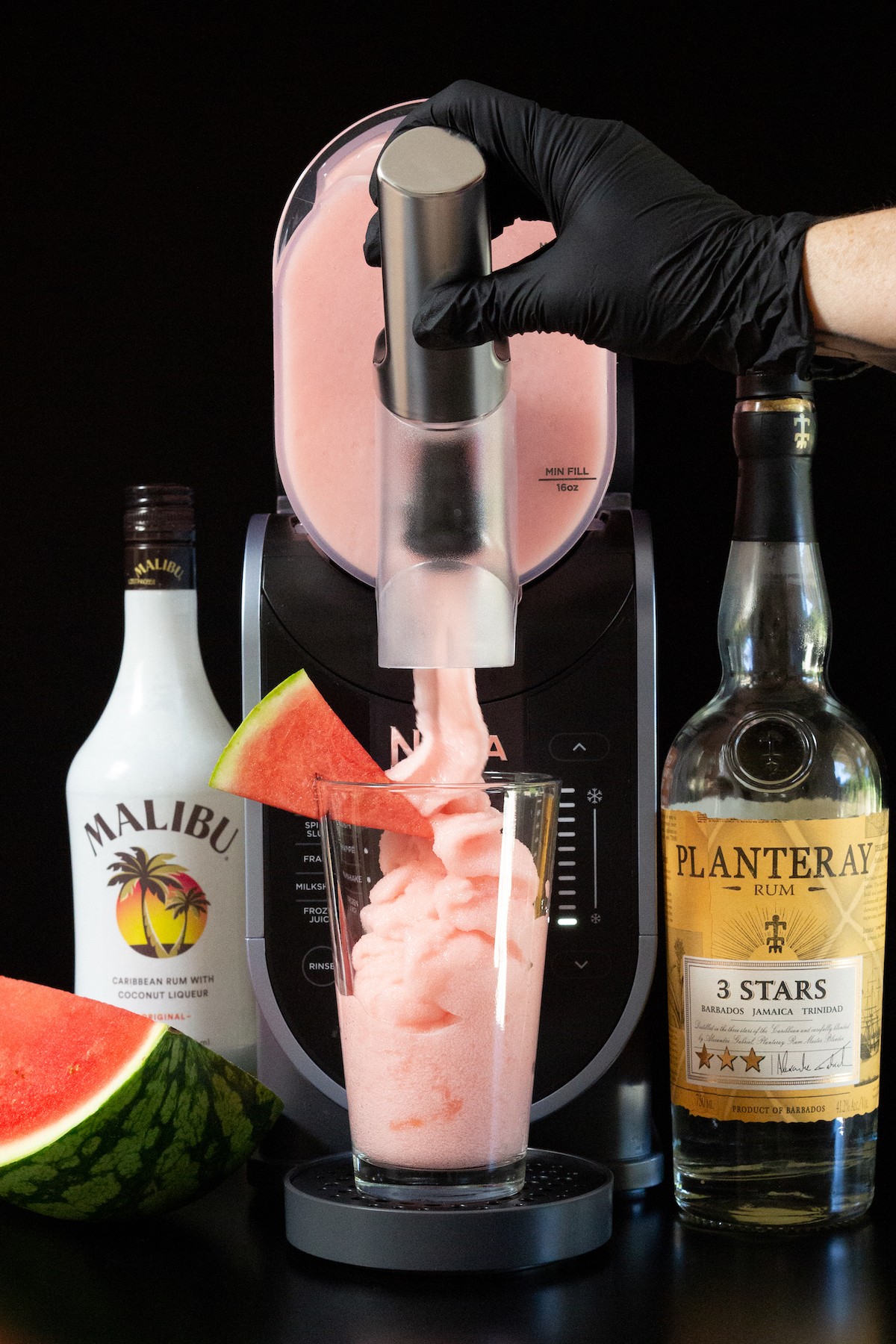 A gloved hand pulls the lever on a Ninja Slushi machine and a pink slushy fills a pint glass that's garnished with a watermelon slice. A bottle of Malibu and a bottle of Jamaican rum are out of focus in the background.
