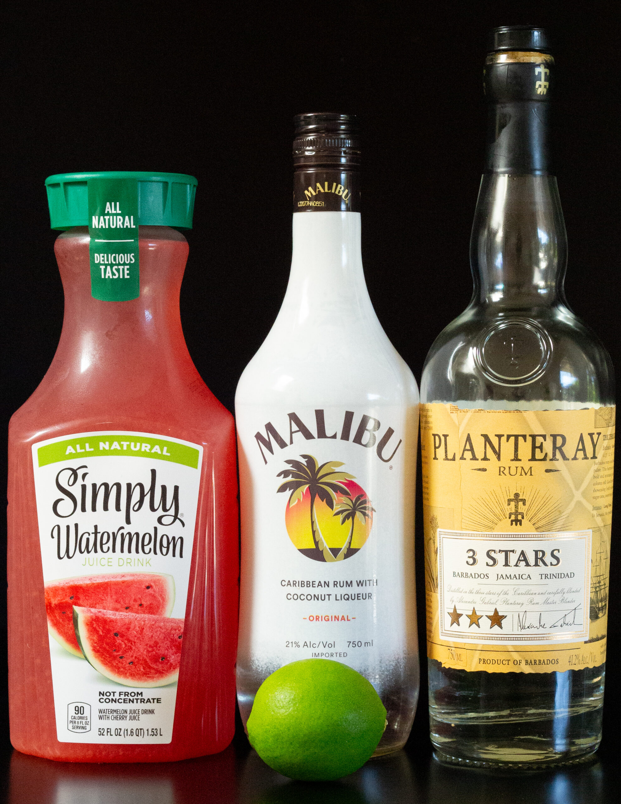 A bottle of Simply Watermelon Juice, Malibu coconut rum, and light Jamaican rum all on a black background.