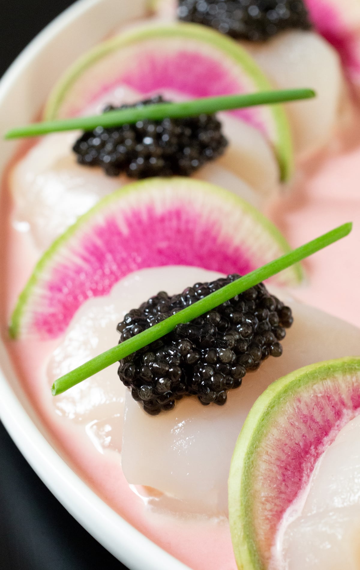 Large white scallops sitting on top of a light pink sauce have been thinly sliced and topped with black caviar and chives. Watermelon radish half-moons rest in-between the scallops.