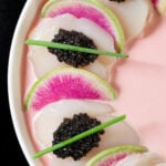 Jumbo large scallops have been sliced thin and stacked accordion style on top of one another. Every other one is garnished with caviar and single chive. Watermelon radishes are placed in between. everything is swimming in a light pink sauce.