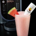 A pint glass filled with a light pink slushy is garnished with a watermelon triangle and a mini bottle of Malibu. A Ninja Slushi machine is out of focus in the background.