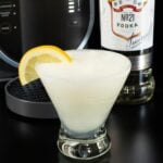 A stemless martini glass filled with a light yellow slushy is garnished with a half lemon moon. A Ninja Slushi machine and a bottle of vodka are out of focus in the background.