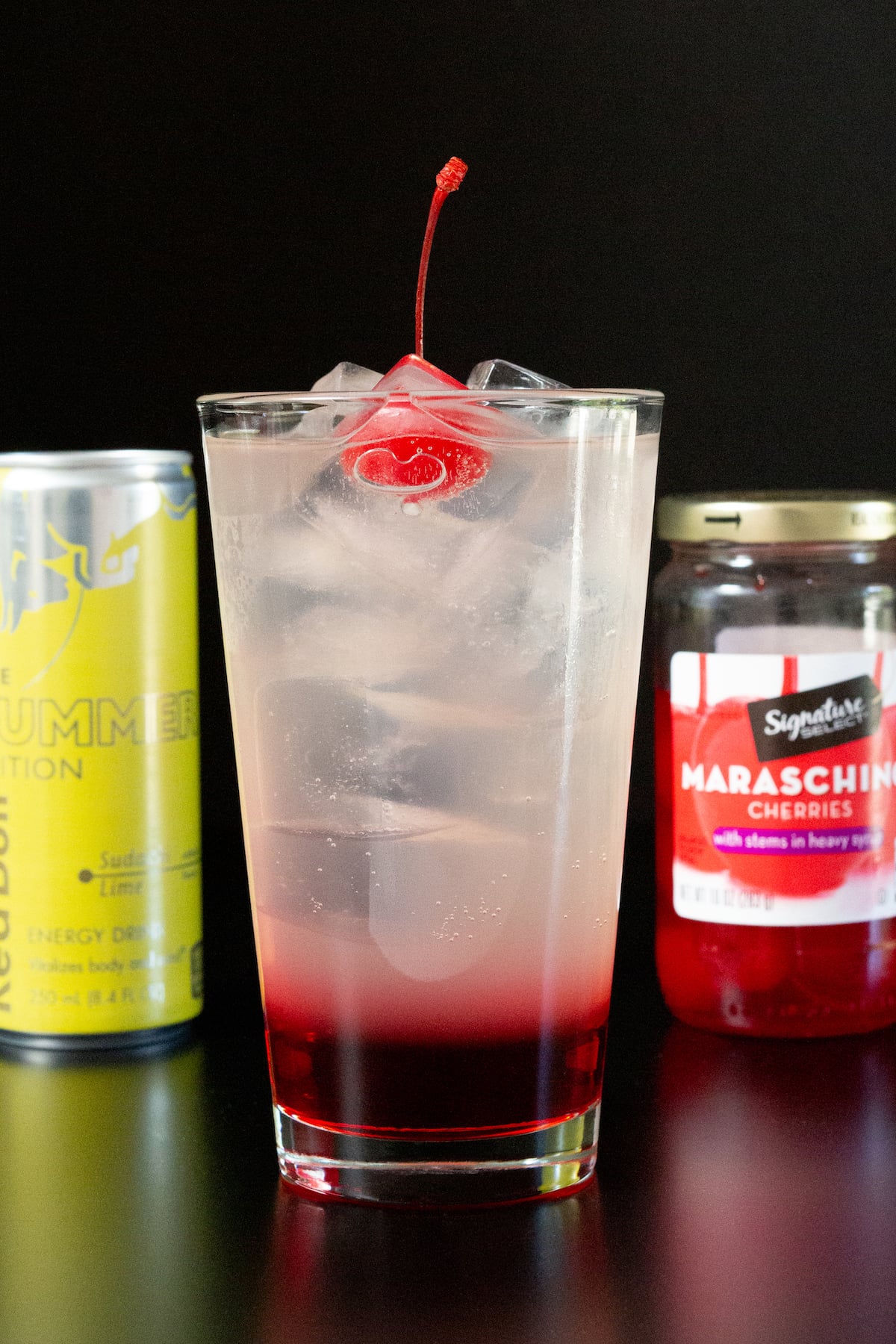 A pint glasss 1/3 filled with a thick, red liquid and the rest of the glass is a translucent liquid. A cherry sits on top. A can of lime Red Bull and a jar of maraschino cherries is out of focus in the background.