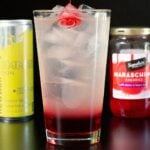 A pint glasss 1/3 filled with a thick, red liquid and the rest of the glass is a translucent liquid. A cherry sits on top. A can of lime Red Bull and a jar of maraschino cherries is out of focus in the background.