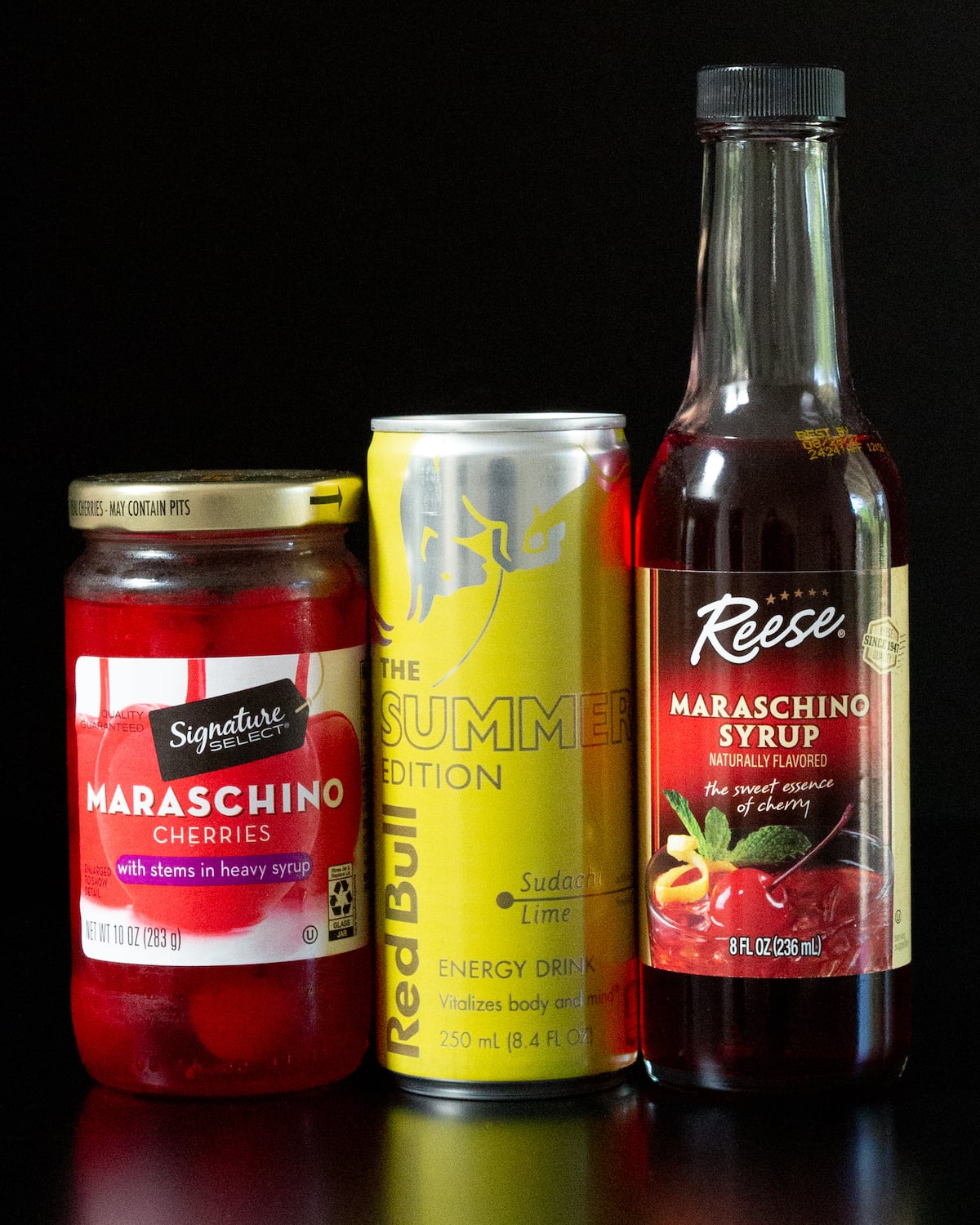 A bottle of maraschino syrup, a can of lime Red Bull, and a bottle of maraschino syrup all on a black background.