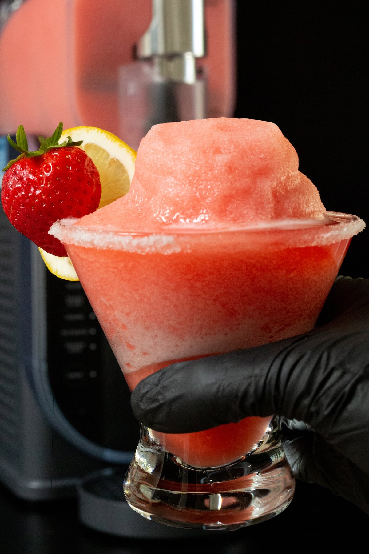 A solved hand holds up a stemless martini glass filled with a light red slushy that's garnished with a lemon wheel and a strawberry. A Ninja Slushi machine is out of focus in the background.