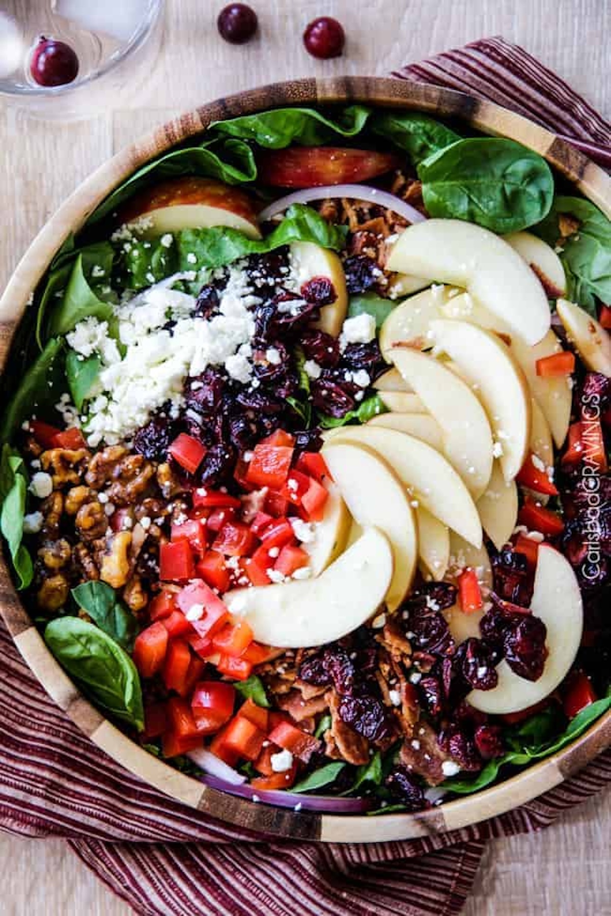 A large wooden salad bowl filled with spinach greens topped with sliced apples, raisins, nuts, crumbled cheese, and diced red pepper.