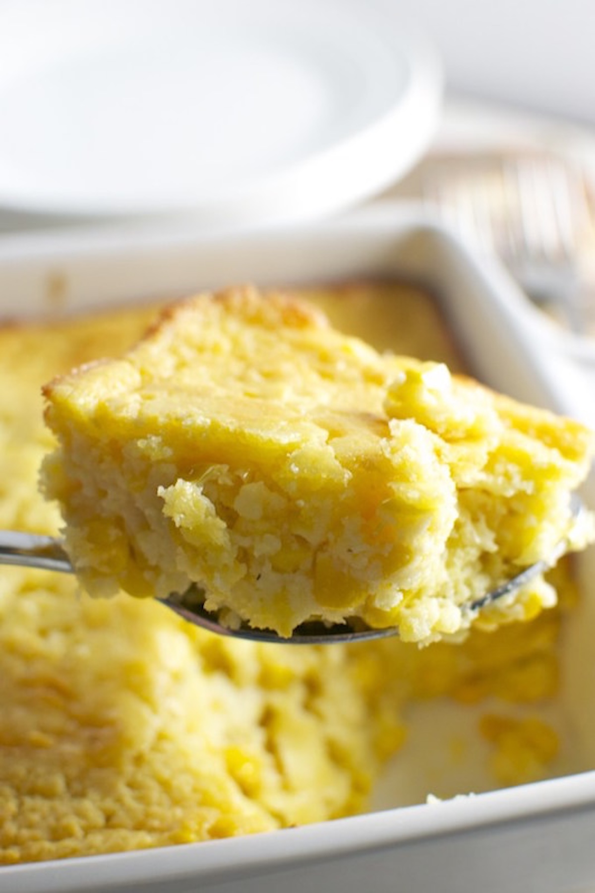 A serving spoon holds up a large square of yellow corn casserole in front of a baking dish with the rest.