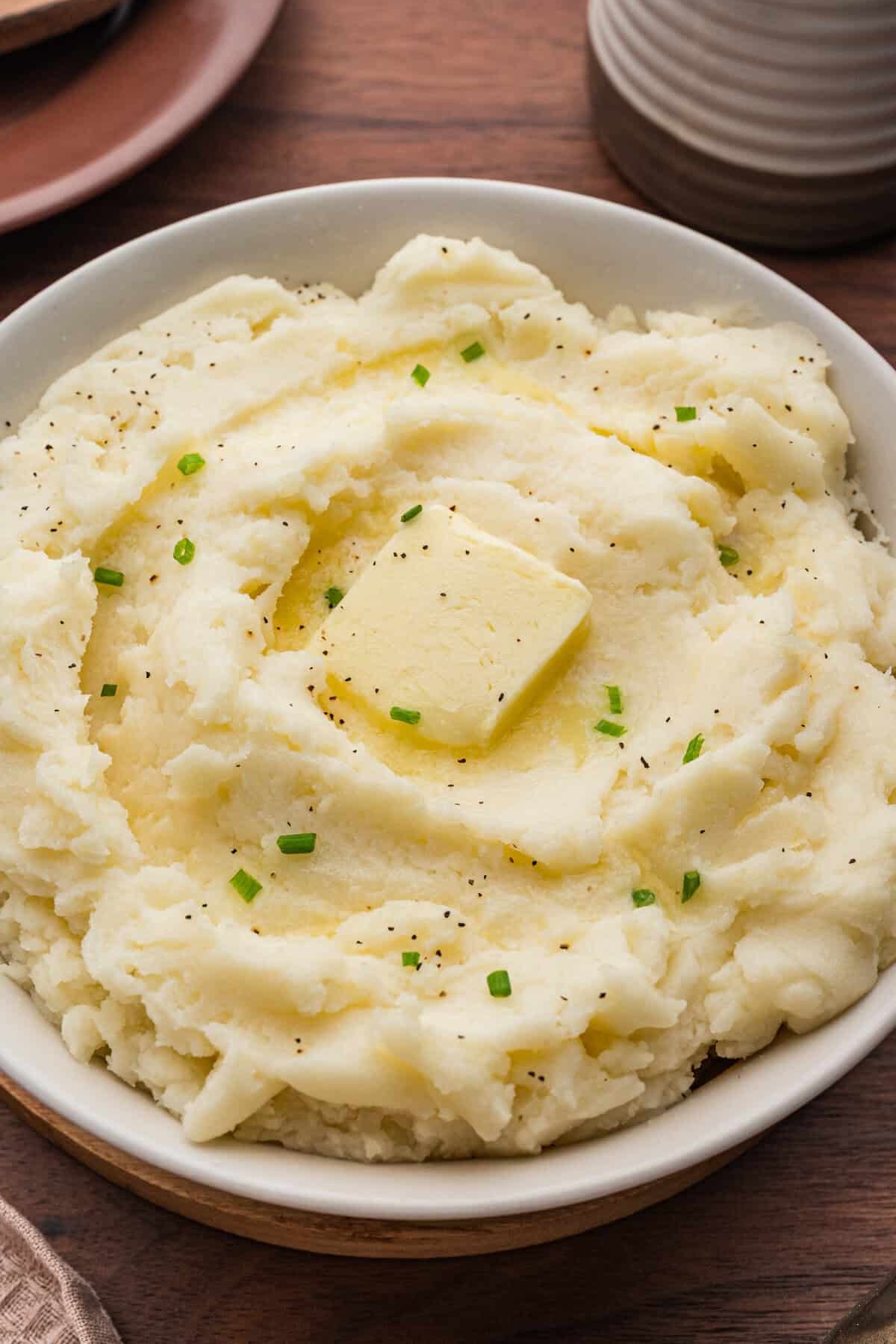 A serving bowl filled with creamy mashed potatoes that have a tablespoon slice of butter in the middle and is lightly garnished with black pepper and chives.
