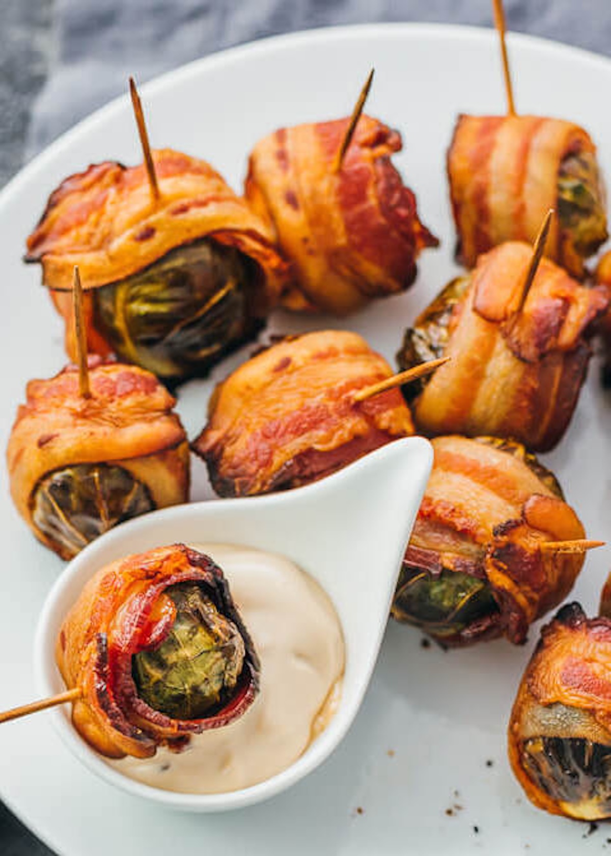 A white serving plate with about a dozen roasted Brussel sprouts wrapped in bacon and stabbed with a toothpick next to a small bowl of dipping sauce.