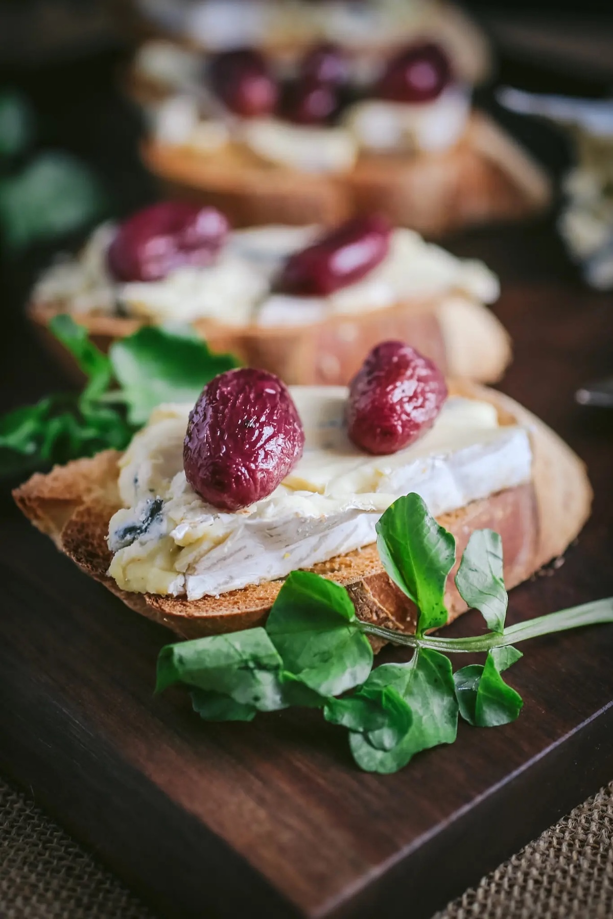 A wood board is filled with crostinis that are topped with thick melted slices of blue cheese and two roasted grapes each.