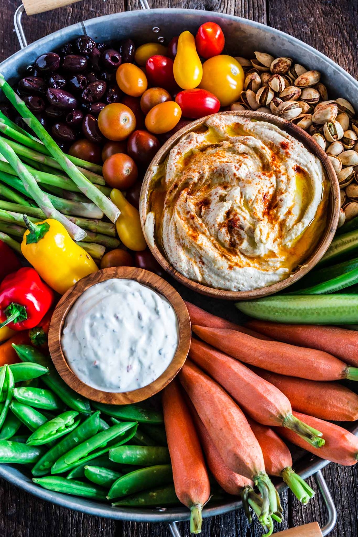 A large metal tray filled with a rustic looking vegetable tray featuring hummus and homemade ranch, pistachios, cherry tomatoes, olives, asparagus, mini peppers, snap peas, carrots, and cucumber.
