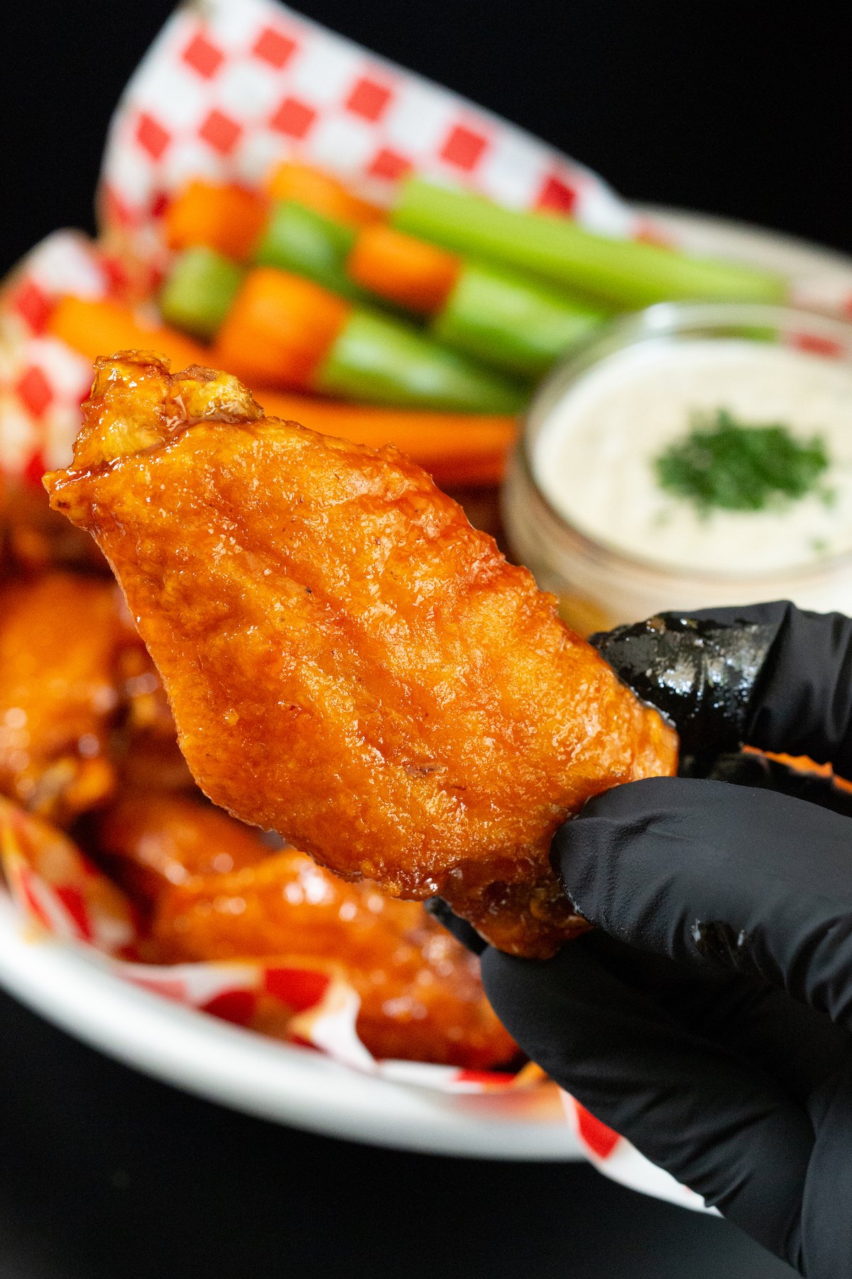 A hand wearing black nitrile gloves holds up a chicken wing coated in hot honey buffalo sauce. A basket of wings is out of focus in the background.