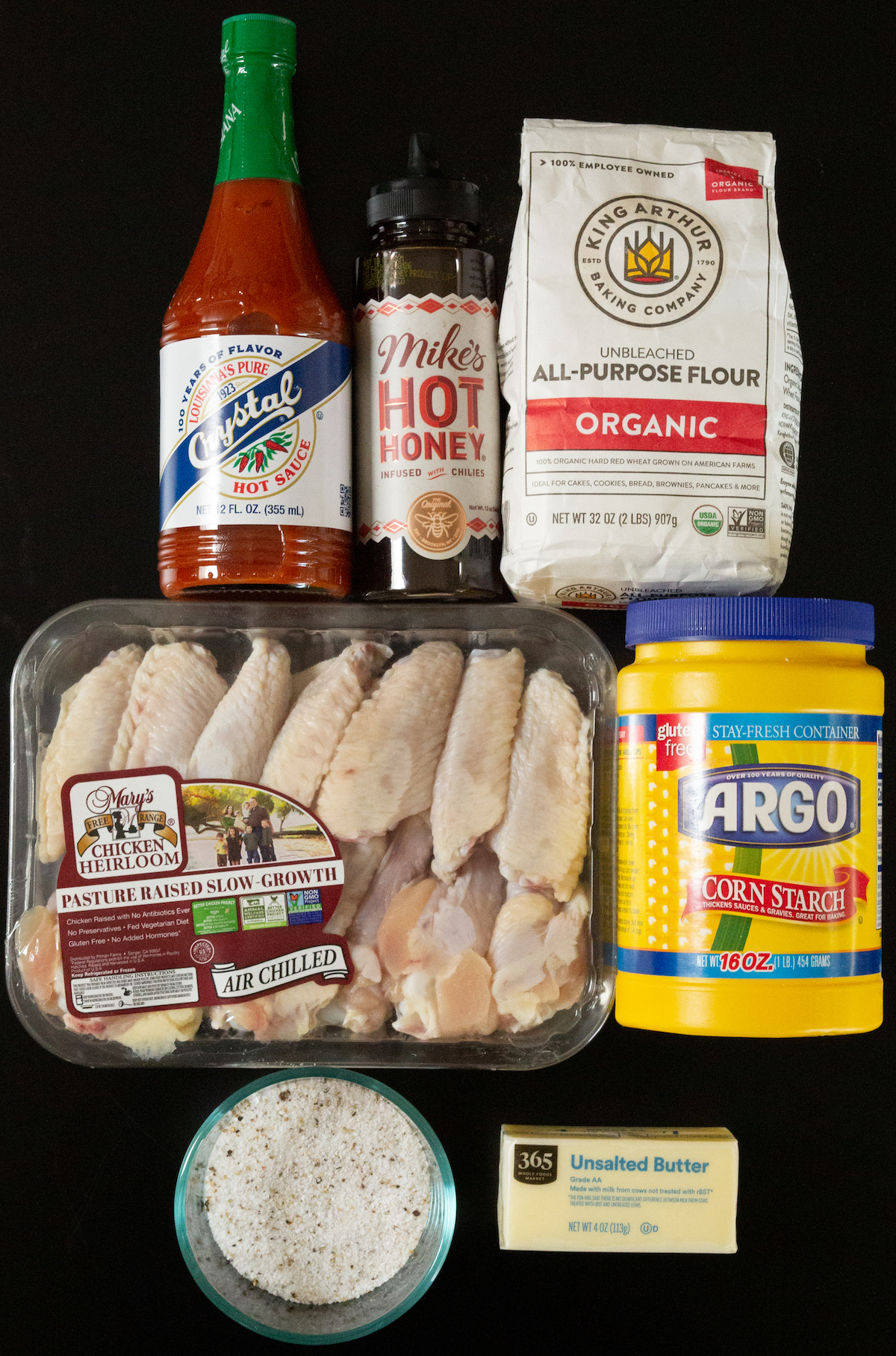 A bottle of Crystal hot sauce, Mike's Hot honey, a bag of all-purpose flour, raw chicken wings, corn starch, and a bowl of seasonings.