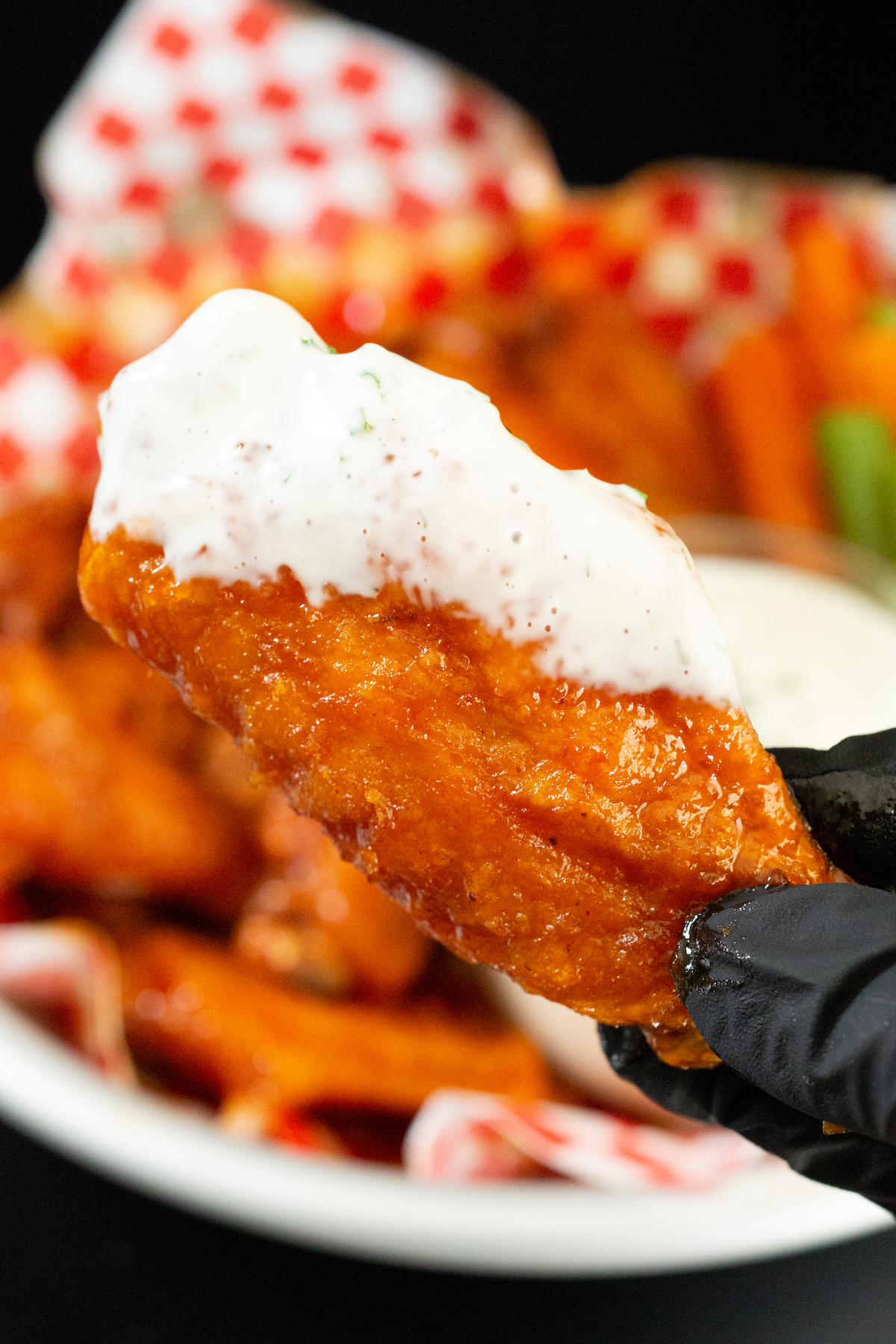A hand wearing black nitrile gloves holds up a hot honey buffalo chicken wing that's been dipped in ranch dressing. A basket of wings is out of focus in the background.