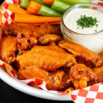 A basket of hot honey buffalo wings with a jar of ranch dressing and celery and carrot sticks.