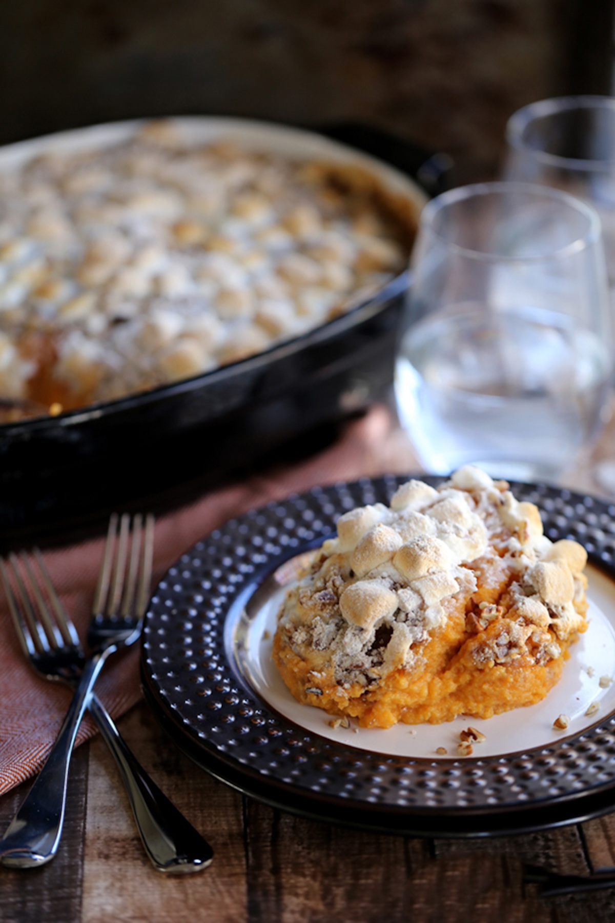 A square of sweet potato casserole topped with toasted marshmallows on a serving plate is in focus in the foreground and a casserole dish with the rest is out of focus in the background.