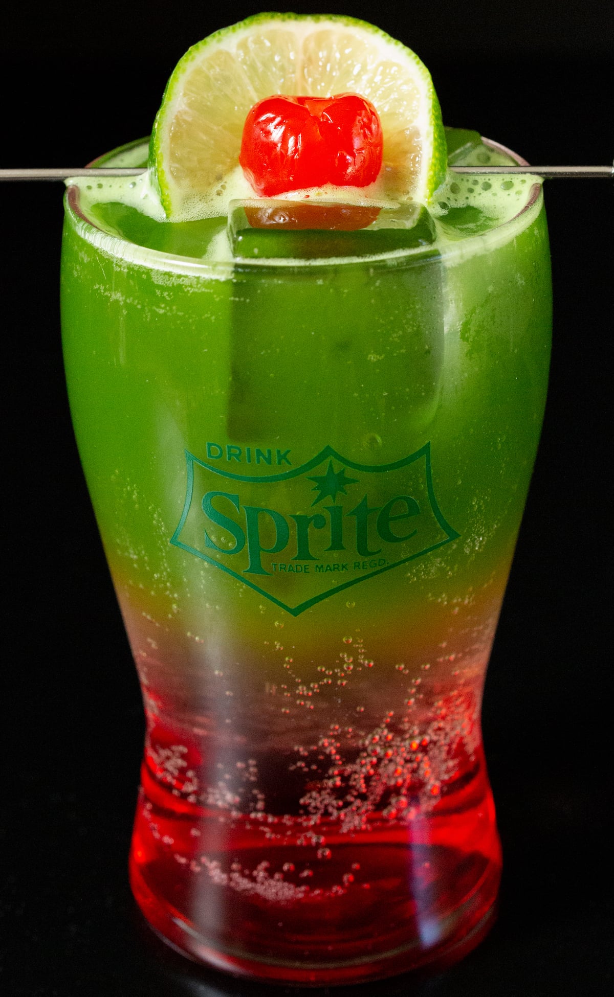 A small hourglass shaped glass is filled halfway with a red Shirley temple mixture and the top half is bright green matcha. It's garnished with a lime wheel and a maraschino cherry.