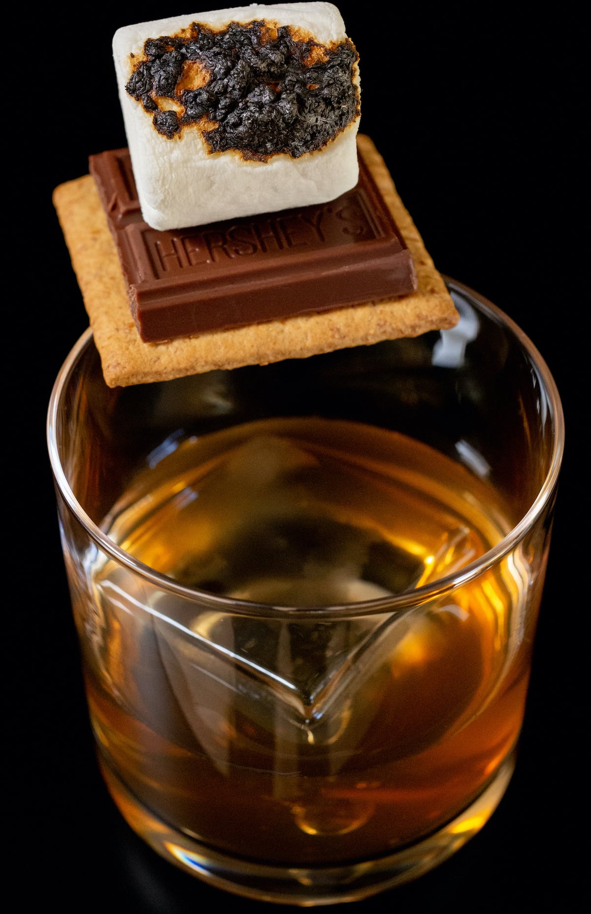 A low ball glass has a large block of clear ice and is about halfway full with an Old Fashioned. A graham cracker, chocolate square, and toasted marshmallow is balanced on the side as a garnish.