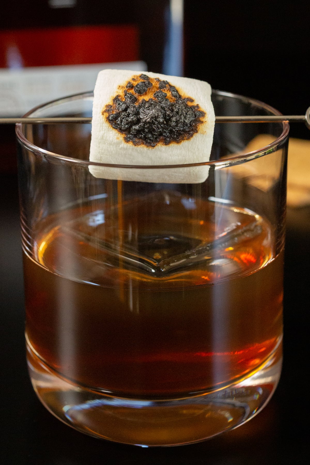 A lowball glass half filled with a reddish orange old fashioned and a large block of ice. A charred marshmallow is skewered across the top.