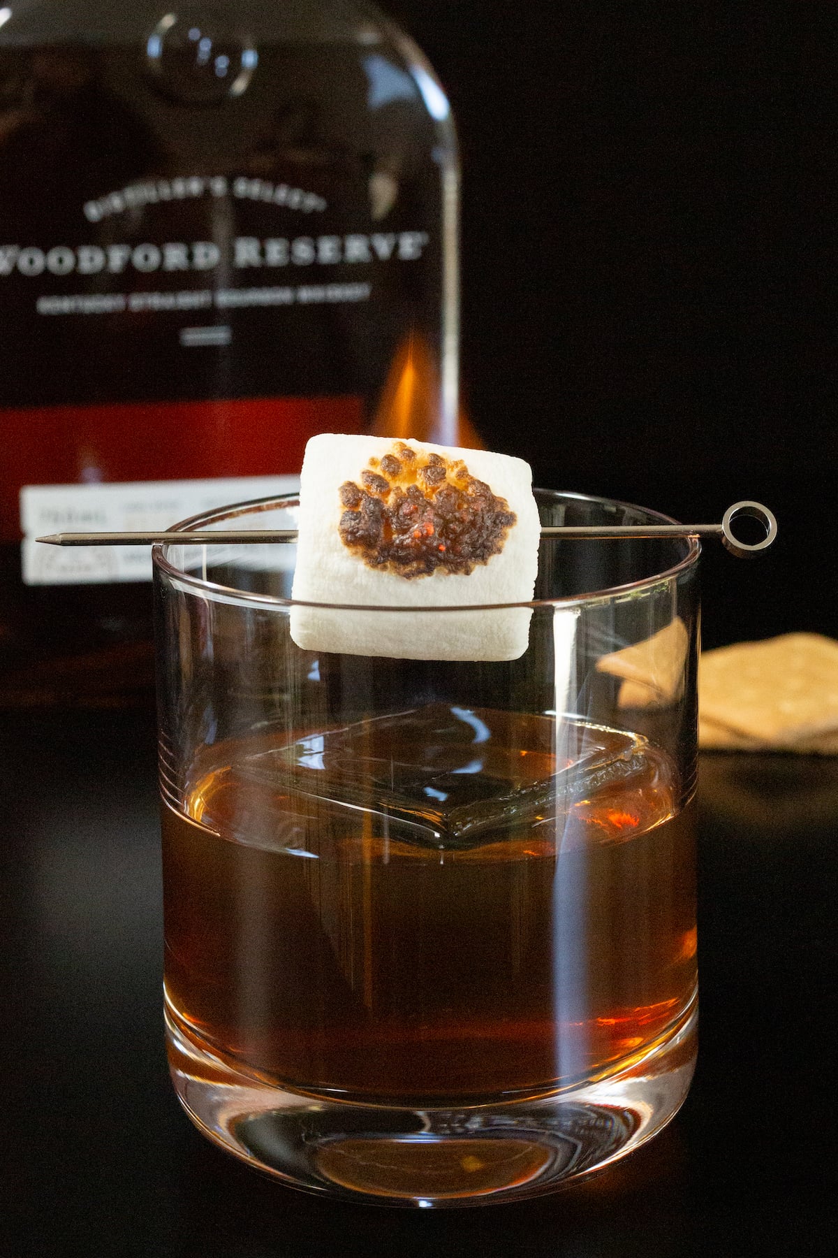 A lowball glass is filled with an old fashioned that has a flaming skewered marshmallow on top. A bottle of Woodford Reserve is out of focus in the background.