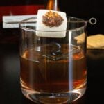 A lowball glass is filled with an old fashioned that has a flaming skewered marshmallow on top. A bottle of Woodford Reserve is out of focus in the background.