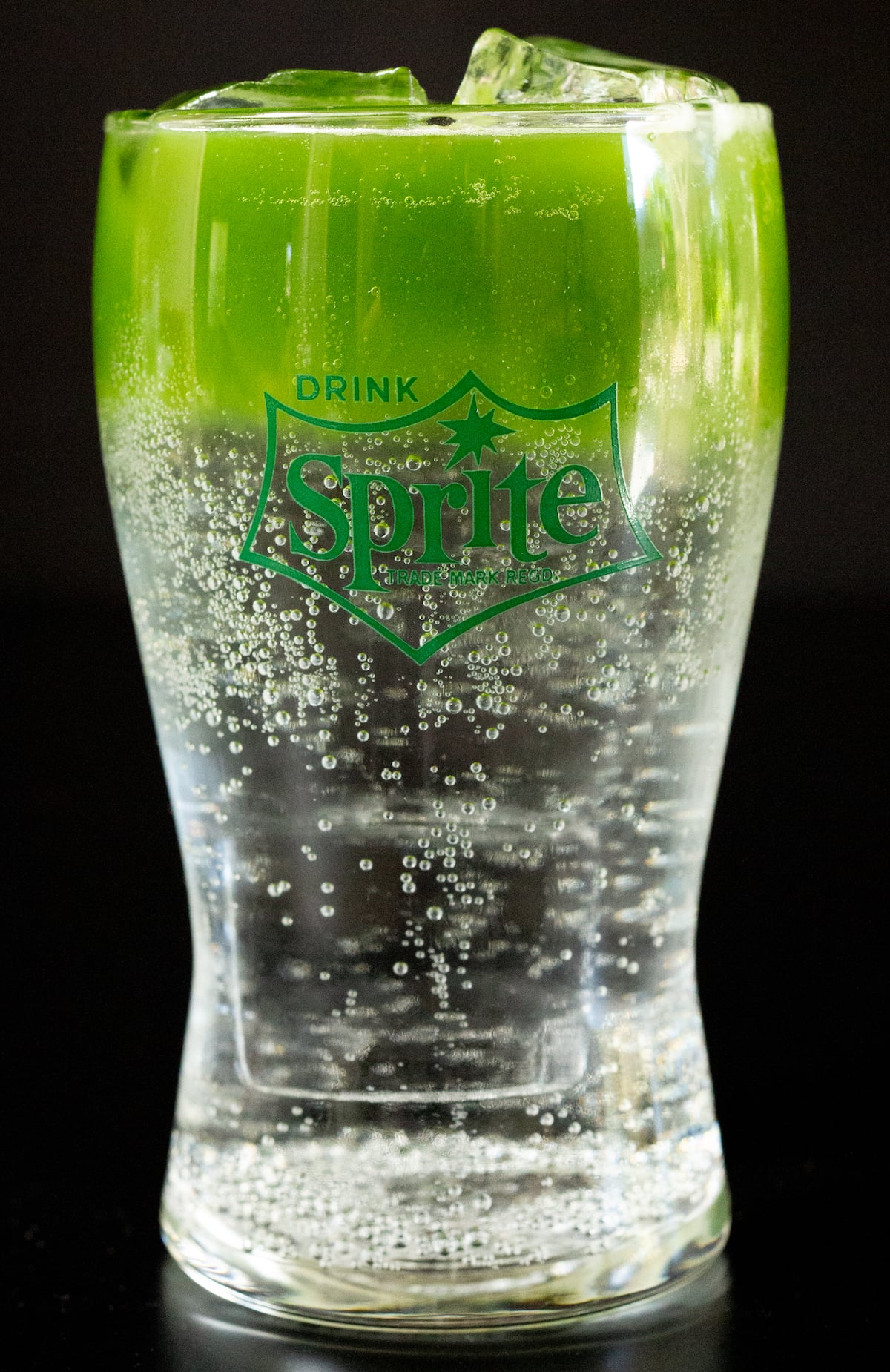 A small hourglass shaped glass is filled 2/3 of the way with Sprite soda and 1/3 with bright green matcha.