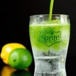 A stream of matcha is being poured into a small glass filled with Sprite and clear ice cubes. A lemon and a lime are out of focus in the background.