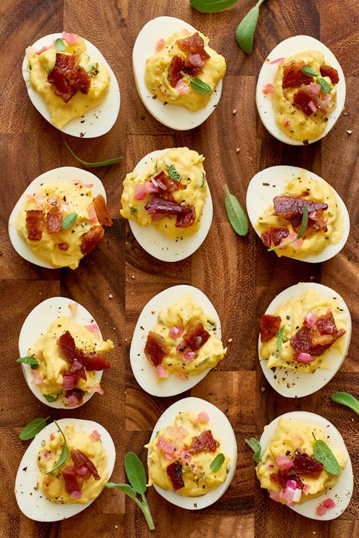 A wooden cutting board with a dozen deviled eggs topped with chopped bacon, shallots, and fresh sage.