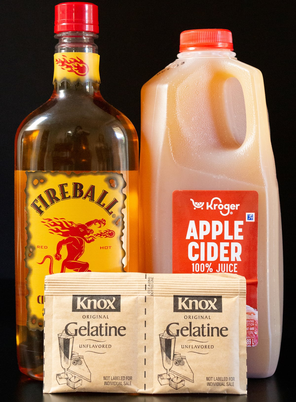 A bottle of Fireball and a gallon of apple cider next to two packets of Knox unflavored gelatin.