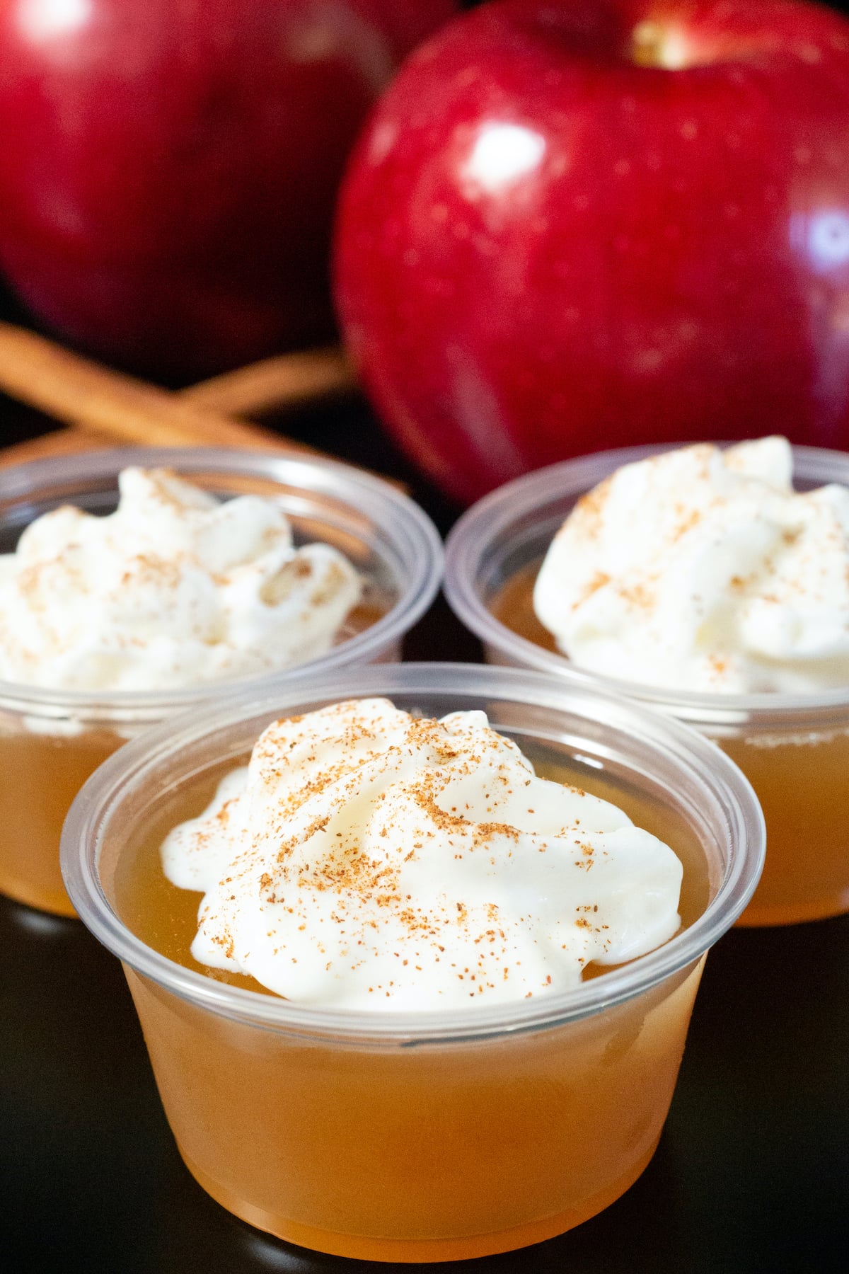 Three plastic 2 ounce cups filled with apple cider jello shots that are topped with a dollop of whipped cream and a pinch of ground cinnamon.