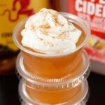 A two ounce plastic cup filled with an apple cider jello shot topped with whipped cream is in focus in the foreground, a bottle of apple cider and Fireball are out of focus in the background.