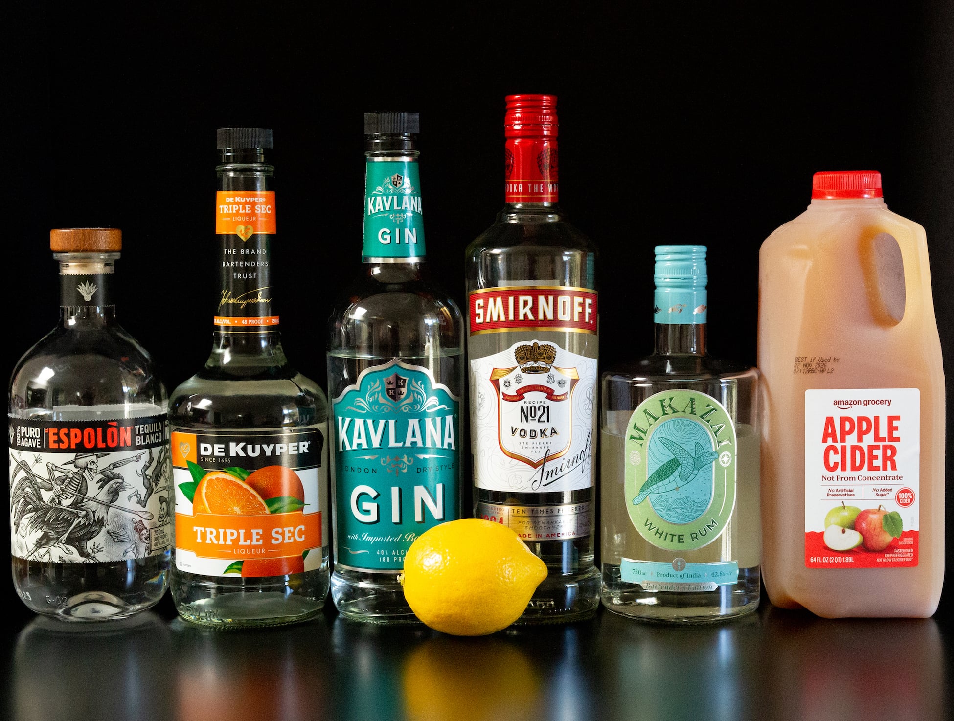 A bottle of blanco tequila, triple sec, gin, vodka, white rum, apple cider, and a whole lemon all on a black background.
