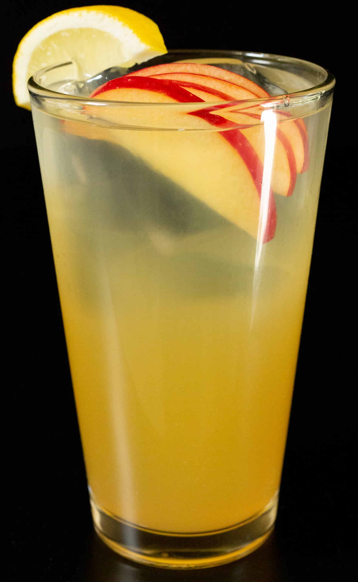 A pint glass is filled with clear ice and a light yellow apple cider long island iced tea cocktail that's garnished with four thin apple sliced inside the glass and a lemon wedge on the rim.
