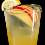 A pint glass is filled with clear ice and a light yellow apple cider long island iced tea cocktail that's garnished with four thin apple sliced inside the glass and a lemon wedge on the rim.
