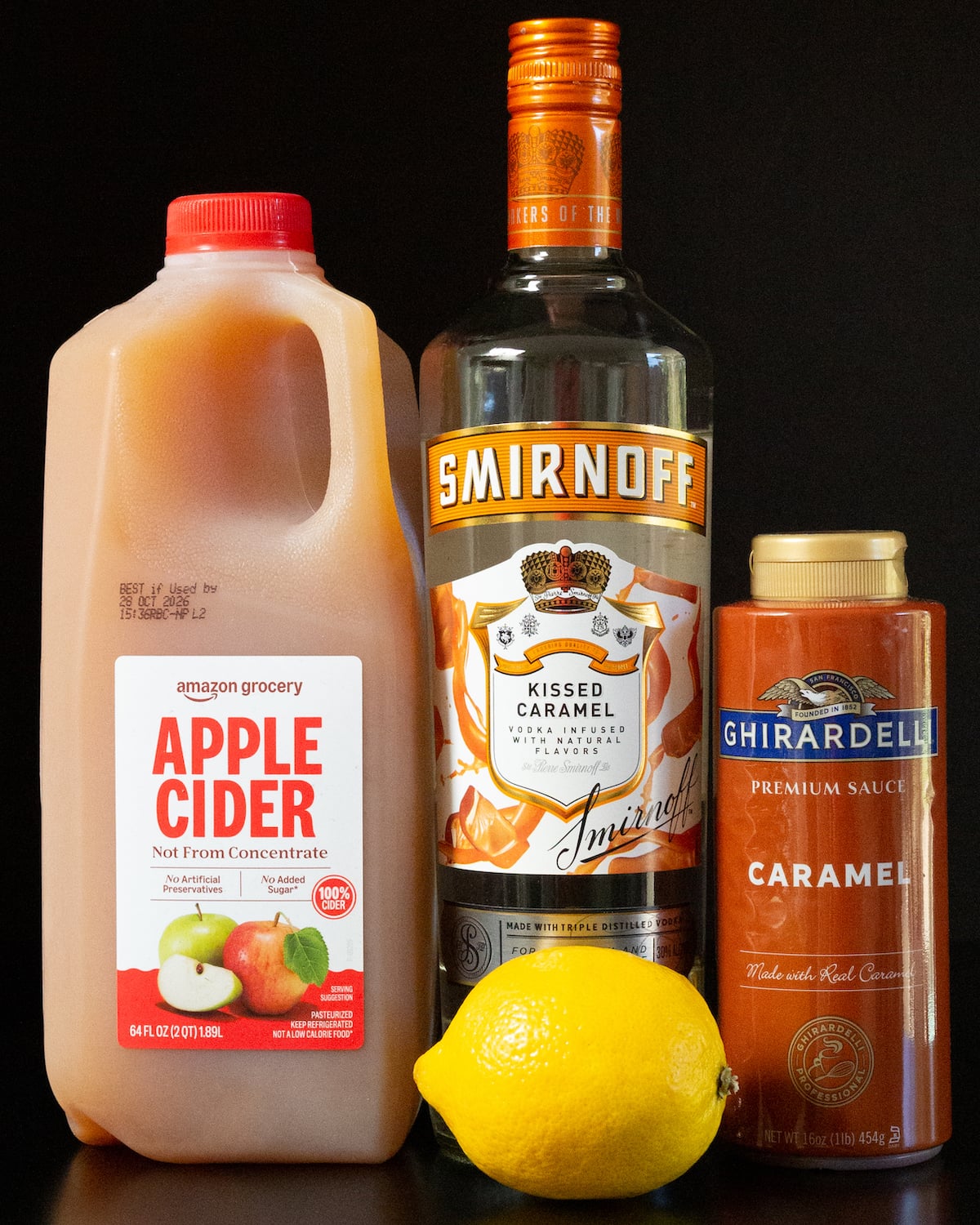 A gallon of apple cider, a bottle of caramel vodka, a bottle of caramel sauce, and a lemon all on a black background.