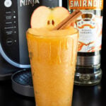 A pint glass filled with an apple cider slushy has caramel dripping down its sides. A bottle of vodka and a Ninja Slushi machine are out of focus in the background.